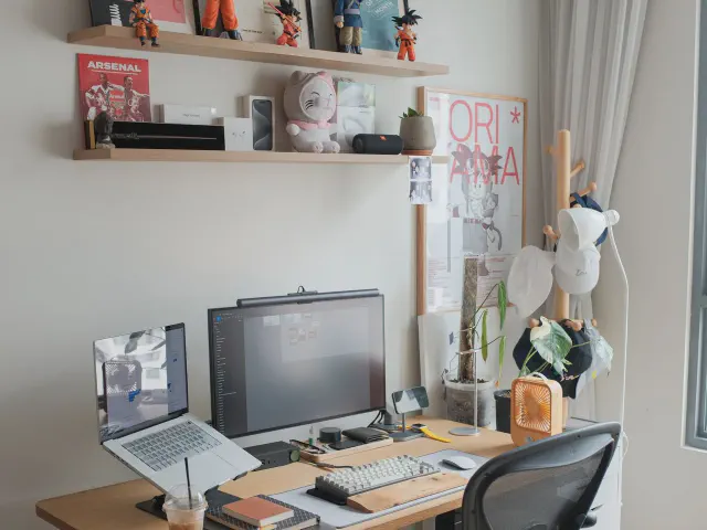 Biurko z dwoma monitorami i laptopem na podstawce, siatkowe krzesło, nad blatem półka z figurkami i kartki przypięte do ściany