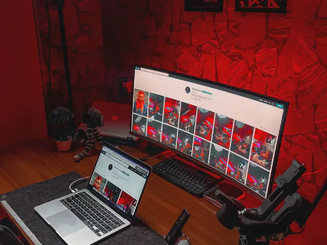 Biurko z zakrzywionym monitorem i laptopem na tle ściany w intensywnym czerwonym oświetleniu, obok statyw z kamerą