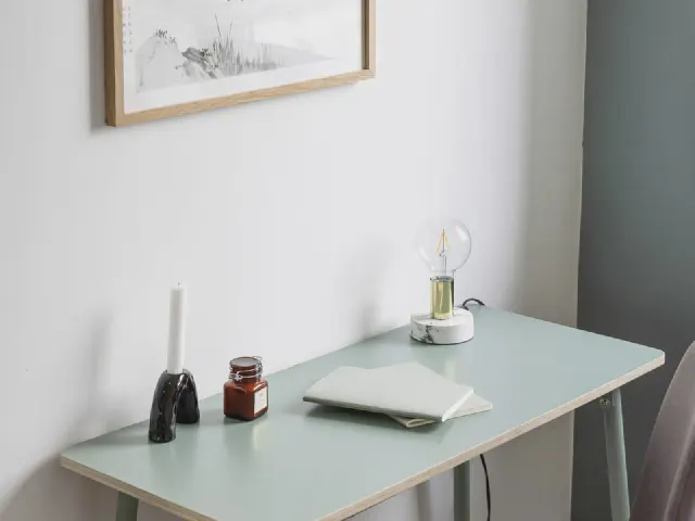 Biurko z miętowym blatem na białych nogach przy białej ścianie, na blacie świeca, słoik, lampka z kloszem i laptop, nad biurkiem grafika w ramie