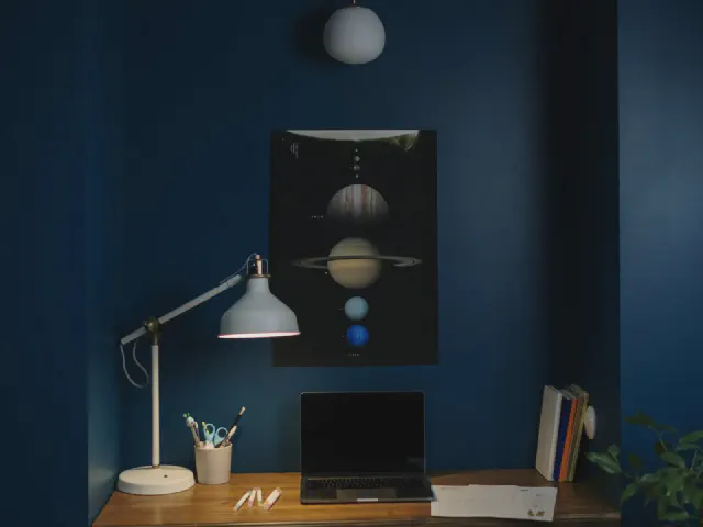 Biurko przy ciemnogranatowej ścianie z plakatem planet, lampa na wysięgniku, laptop i przybory w kubku