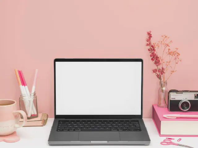 Biurko przy pudroworóżowej ścianie, laptop, różowe pudełka z aparatem, kubek, przybory w słoiku i suszone kwiaty w wazonie