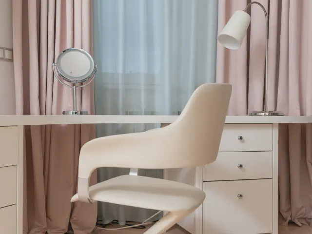 Białe biurko z szufladami przy oknie z pudroworóżowymi zasłonami i firaną, kremowe krzesło, lusterko i lampa podłogowa