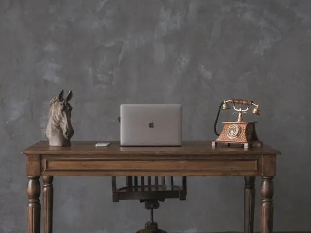 Ciemnoszara ściana z efektem przecieranego tynku, drewniane biurko na toczonych nogach, laptop, figurka konia i telefon w stylu retro