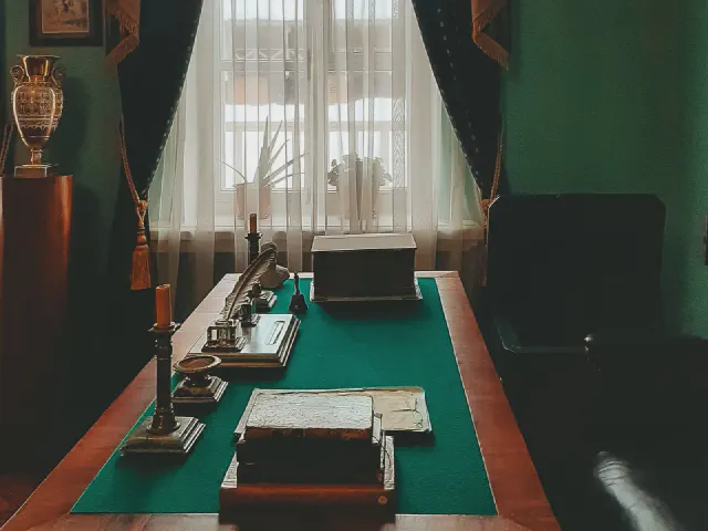 Gabinet z zielonymi ścianami i ciemnymi zasłonami, biurko z blatem obitym zieloną skórą, lampa, przybory i dokumenty na blacie, w tle okno z firaną