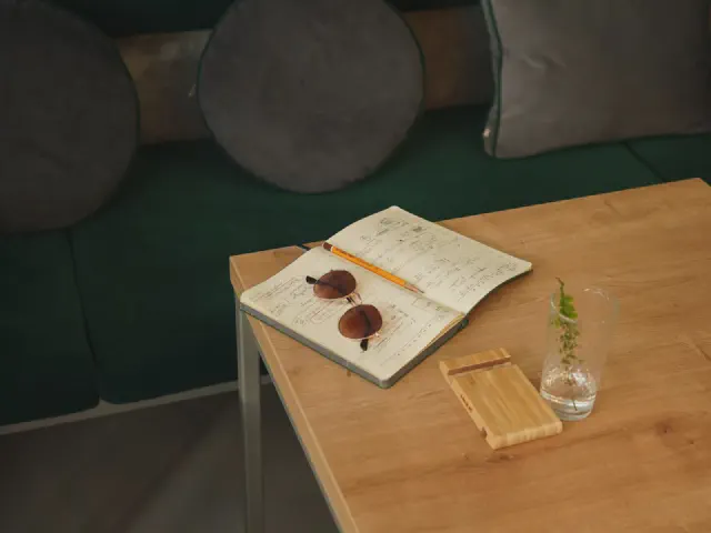 Drewniany stolik z otwartym notesem, okularami i małym wazonem przy ciemnozielonej welurowej sofie