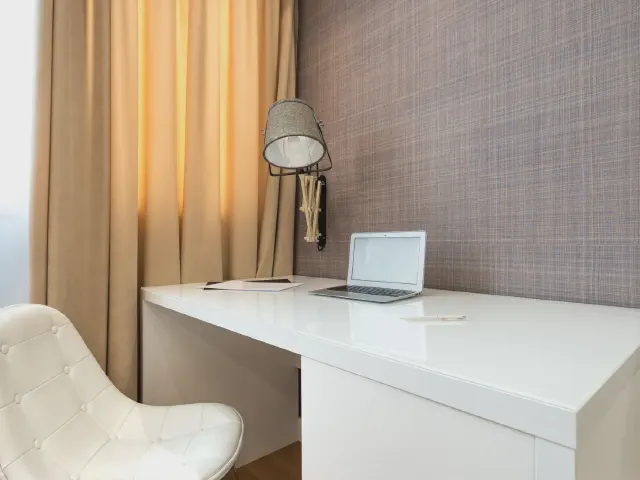Białe biurko z lakierowanym blatem pod oknem z beżową zasłoną, laptop, lampka na wysięgniku przy ścianie z fakturą i biały pikowany fotel