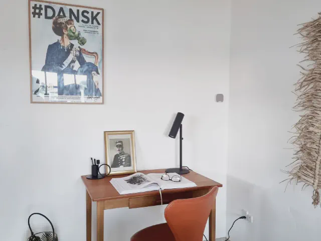 Małe drewniane biurko przy białej ścianie, na nim czarna lampka i grafika w ramce, nad biurkiem plakat, obok pomarańczowe krzesło i suszone trawy
