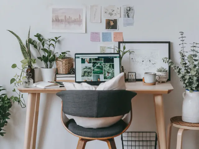 Biurko z jasnego drewna z monitorem, ciemne krzesło z giętą drewnianą ramą, dużo roślin w doniczkach i kartki z inspiracjami przypięte do białej ściany