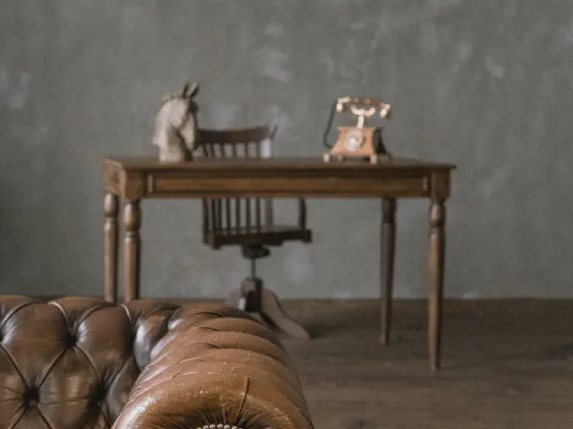 Stylowe drewniane biurko na toczonych nogach z krzesłem obrotowym, na blacie figurka konia i stary telefon, na pierwszym planie brązowa skórzana sofa chesterfield
