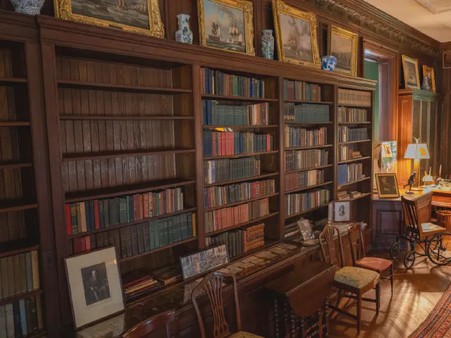 Domowa biblioteka z regałami z ciemnego drewna od podłogi po sufit wypełnionymi książkami, obrazy w złotych ramach nad regałami i drewniane krzesła