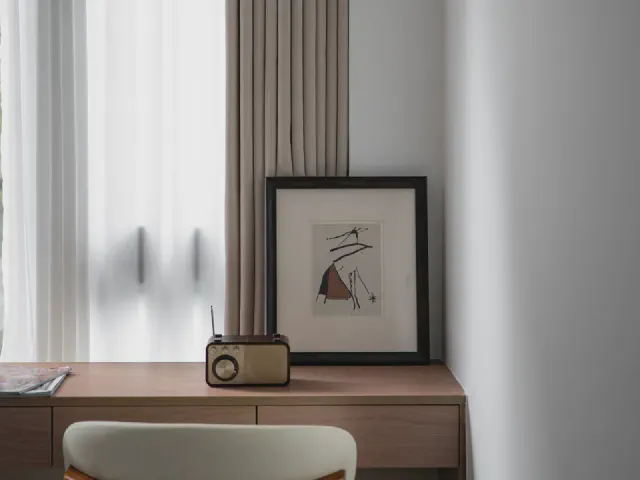Kącik przy oknie z niskim drewnianym blatem, radiem w stylu retro, grafiką w czarnej ramie opartą o ścianę i szarą zasłoną