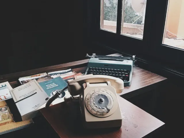 Ciemne drewniane biurko przy oknie, beżowy telefon z tarczą, turkusowa maszyna do pisania, książki i notatki na blacie