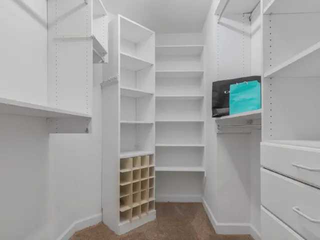 Mała garderoba typu walk-in z białymi półkami, drążkami, szufladami i przegródkami na buty, na podłodze wykładzina