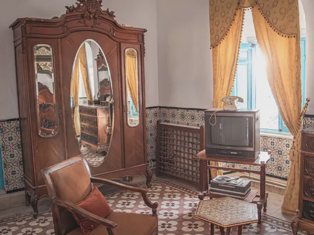 Stara drewniana szafa z trzema lustrami, obok retro telewizor na stoliku, fotel i ścienne płytki w ornament