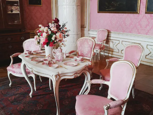 Stylowa jadalnia z różowymi ścianami w ornament, rzeźbionym białym stołem i krzesłami obitymi różowym aksamitem, na stole bukiet i porcelana