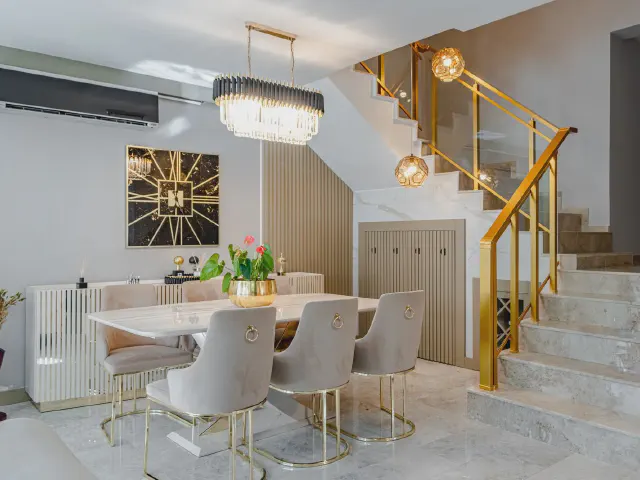 Jadalnia glamour przy schodach ze złotą balustradą – biały stół z szarymi welurowymi krzesłami na złotych nogach, kryształowy żyrandol i marmurowa podłoga