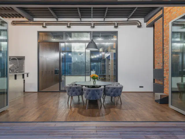 Loft z drewnianą podłogą i przeszkloną ścianą – okrągły stół z szarymi pikowanymi fotelami pod czarną lampą, w tle ściana z czerwonej cegły i stalowe belki pod sufitem