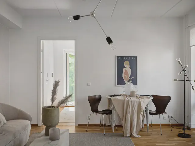 Okrągły stół nakryty lnianym obrusem z ciemnymi krzesłami ze sklejki, nad nim czarna lampa z ramionami, plakat na białej ścianie i szara sofa