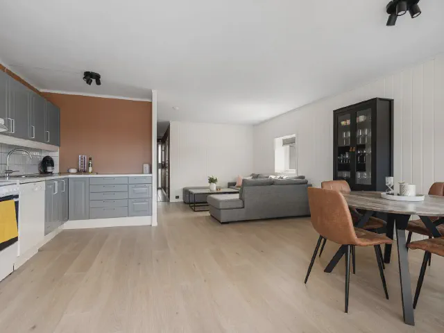 Kuchnia w układzie L z szarymi frontami otwarta na salon, drewniany stół z brązowymi krzesłami i szara sofa w głębi