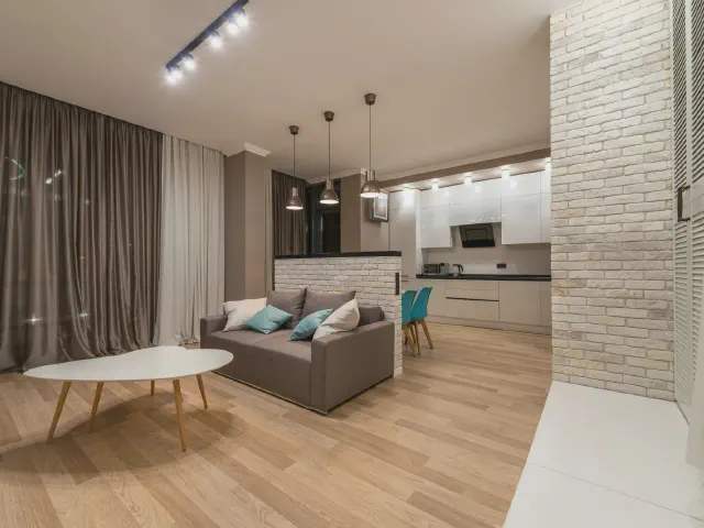 Otwarta kuchnia oddzielona od salonu niską ścianką z białej cegły z blatem, przed nią brązowoszara sofa i owalny biały stolik