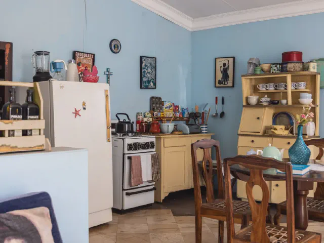 Kuchnia z błękitnymi ścianami, kremowym kredensem pełnym filiżanek, starą kuchenką gazową i drewnianym stołem z krzesłami