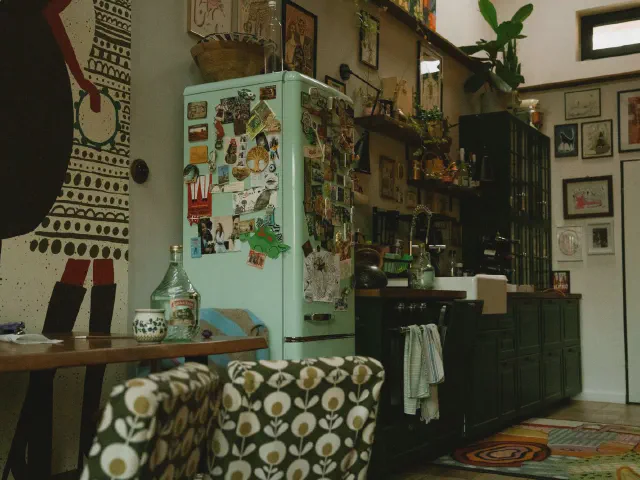 Miętowa lodówka retro pokryta magnesami, zielone szafki, mural na ścianie i krzesła we wzór w kwiaty