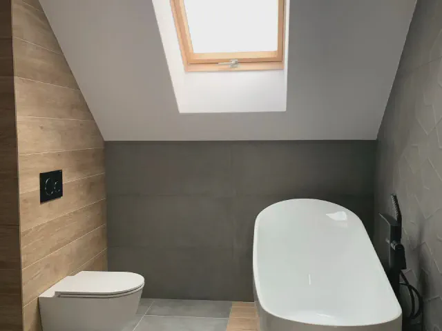 Łazienka na poddaszu z oknem dachowym w skosie, wanną wolnostojącą ustawioną pod skosem, wiszącą toaletą i ścianą obłożoną drewnopodobnymi płytkami
