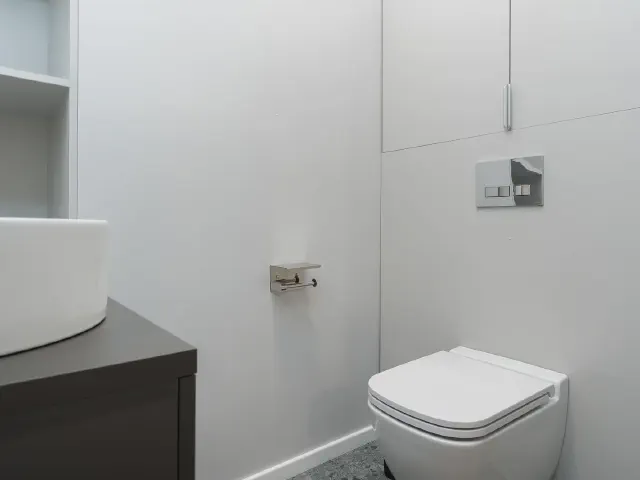 Białe WC z wiszącą miską, przyciskiem spłukiwania na zabudowie stelaża, uchwytem na papier i umywalką na ciemnej szafce