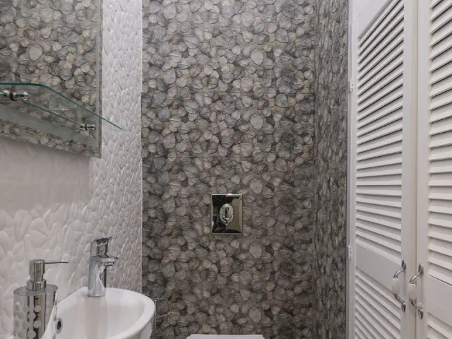 Wąskie WC z mozaiką w szarym kamiennym wzorze na ścianie za stelażem, małą umywalką i żaluzjowymi drzwiami