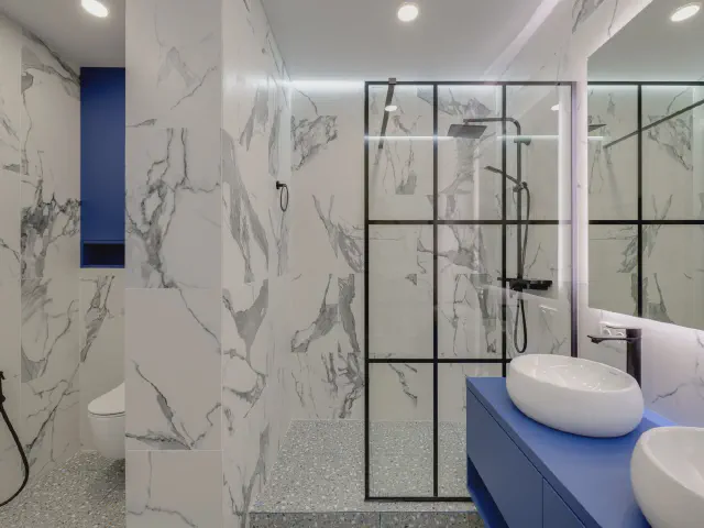 Prysznic walk-in za szklaną ścianką z czarną siatką profili, ściany w płytkach w rysunku marmuru i niebieska szafka z umywalką nablatową