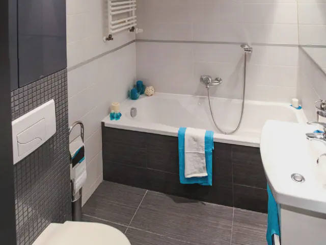 Wanna z ciemną obudową w jasnej łazience, nad nią bateria z prysznicem i ściana z pasem mozaiki