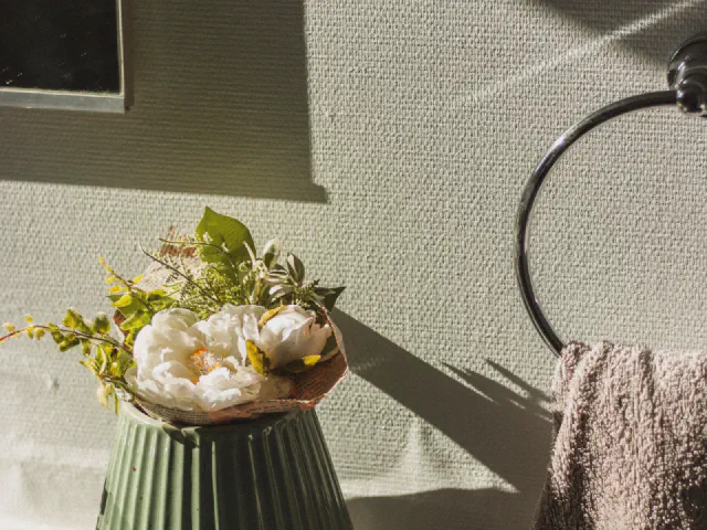 Zielony ceramiczny wazon z kwiatami przy umywalce na tle ściany w szarozielonym odcieniu, obok ręcznik na okrągłym wieszaku