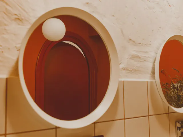 Ściana w kolorze terakoty z owalnymi lustrami w grubych białych ramach, kulistym kinkietem i jasnymi płytkami do połowy wysokości