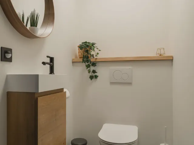 Mała toaleta z podwieszaną miską WC, szafką z drewnianym frontem, okrągłym lustrem w drewnianej ramie, półką z rośliną i posadzką we wzór sześcianów
