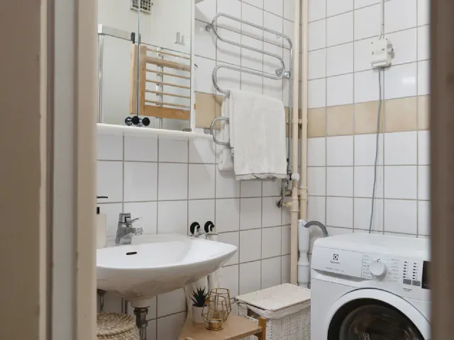 Mała łazienka z białymi kwadratowymi płytkami, umywalką, grzejnikiem na ręczniki, pralką i drewnianym regałem z koszykami