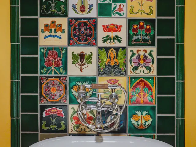 Kolorowe płytki z ornamentami roślinnymi otoczone zielonymi kaflami nad umywalką z chromowaną baterią retro