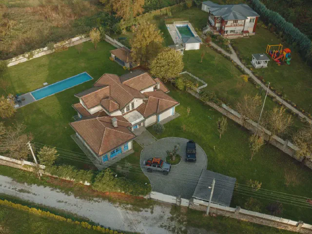 Widok z drona na dużą działkę z domem, podjazdem, prostokątnym basenem, placem zabaw i zadrzewioną granicą