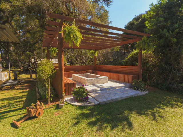 Drewniana pergola na trawniku nad kamiennym podestem z wbudowanymi ławkami i paleniskiem, obok zwisające paprocie w donicach