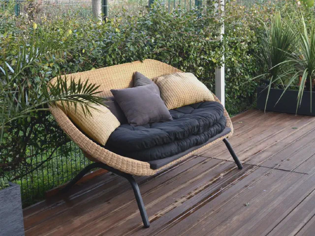 Rattanowa sofa z granatowym siedziskiem i beżowymi poduszkami na tarasie z desek, obok gęste krzewy i trawy w donicach