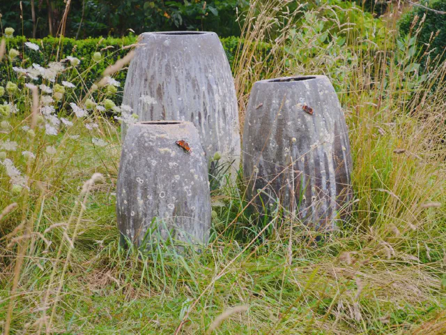 Trzy wysokie, szare donice ceramiczne ustawione w wysokich trawach ozdobnych