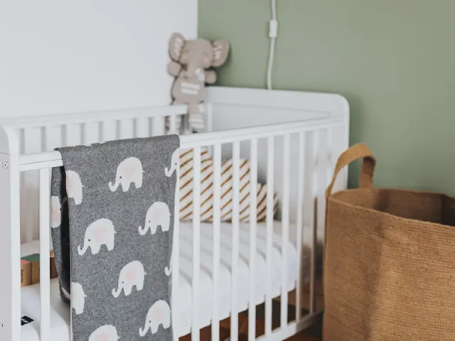 Białe łóżeczko z szarym kocykiem w słonie przy szałwiowozielonej ścianie, pluszowy słonik i jutowy kosz obok
