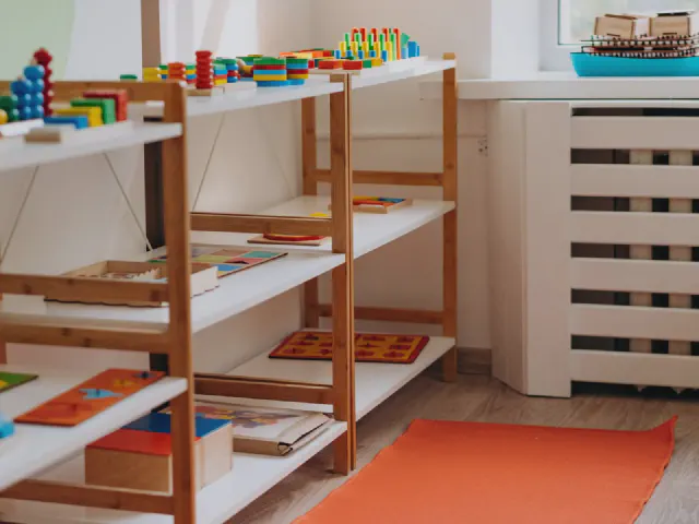 Niskie drewniane regały z białymi półkami ustawione przy ścianie, na nich poukładane zabawki edukacyjne, pod spodem pomarańczowy dywanik