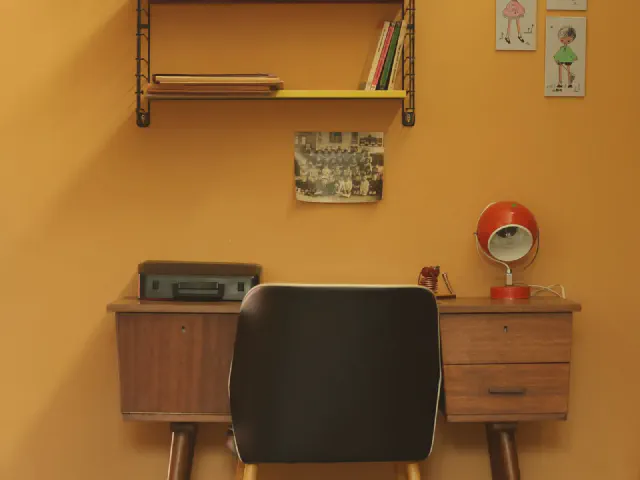 Drewniane biurko w stylu retro z szufladami przy pomarańczowej ścianie, czarne krzesło, czerwona lampka, półka z książkami i obrazki nad blatem