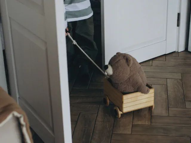 Pluszowy miś w drewnianym wózku na podłodze z brązowego parkietu w jodełkę, obok szare drzwi