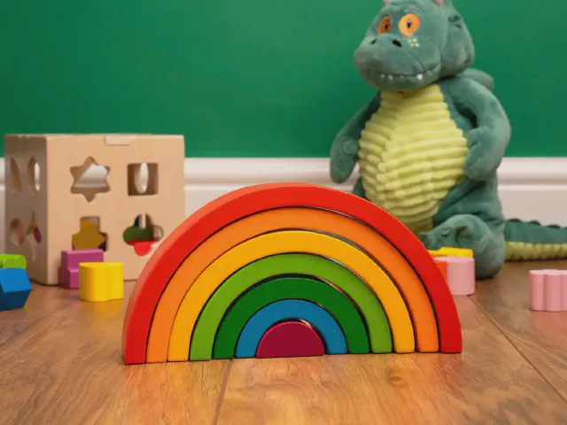 Drewniana tęcza do układania, sorter z klockami i pluszowy zielony dinozaur na drewnianej podłodze przy ciemnozielonej ścianie