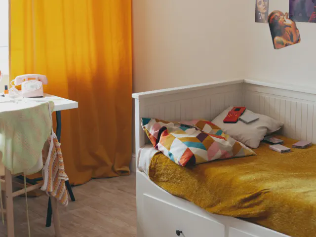 Pokój dziecięcy z białym łóżkiem przykrytym musztardową narzutą i kolorowymi poduszkami, musztardowymi zasłonami przy oknie i białym biurkiem z krzesłem