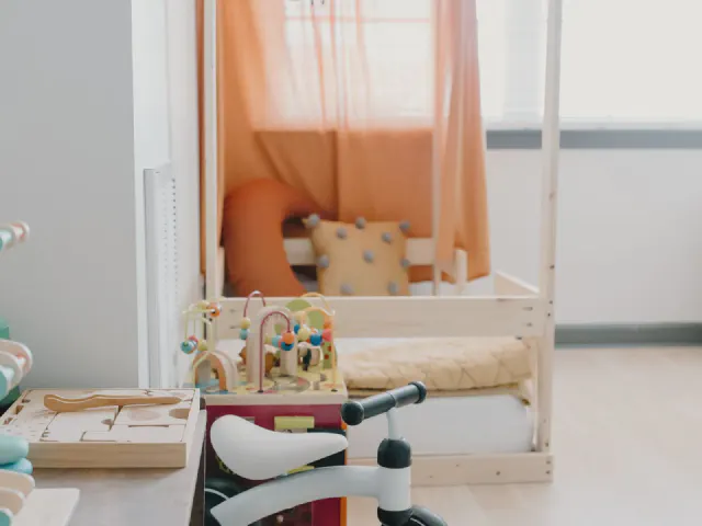 Łóżko domek z jasnego drewna z pomarańczową zasłoną, drewniane zabawki na niskiej półce i biały rowerek biegowy w jasnym pokoju