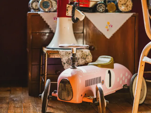 Różowy samochodzik na pedały w stylu retro obok drewnianej komody z haftowaną serwetką i lampą z ceramiczną podstawą
