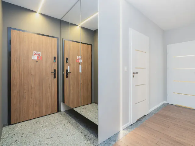 Przedpokój z lustrzaną ścianą w szarej wnęce odbijającą drewniane drzwi wejściowe, obok białe drzwi i posadzka z lastryko