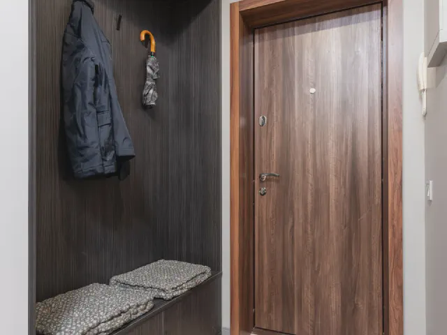 Wnęka z ciemnego drewna z haczykami na kurtki i parasol oraz siedziskiem z miękkimi poduszkami, obok drewniane drzwi wejściowe
